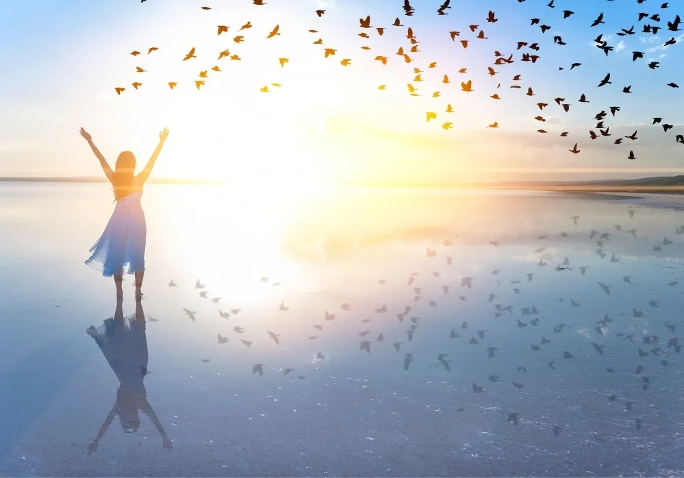 Woman standing on the shore at sunrise, arms raised, with birds flying in the sky – symbolizing freedom and inner peace.