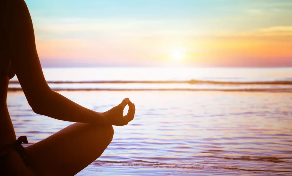 Person practicing Sukhasana yoga pose at sunrise by the beach promoting relaxation and mindfulness.