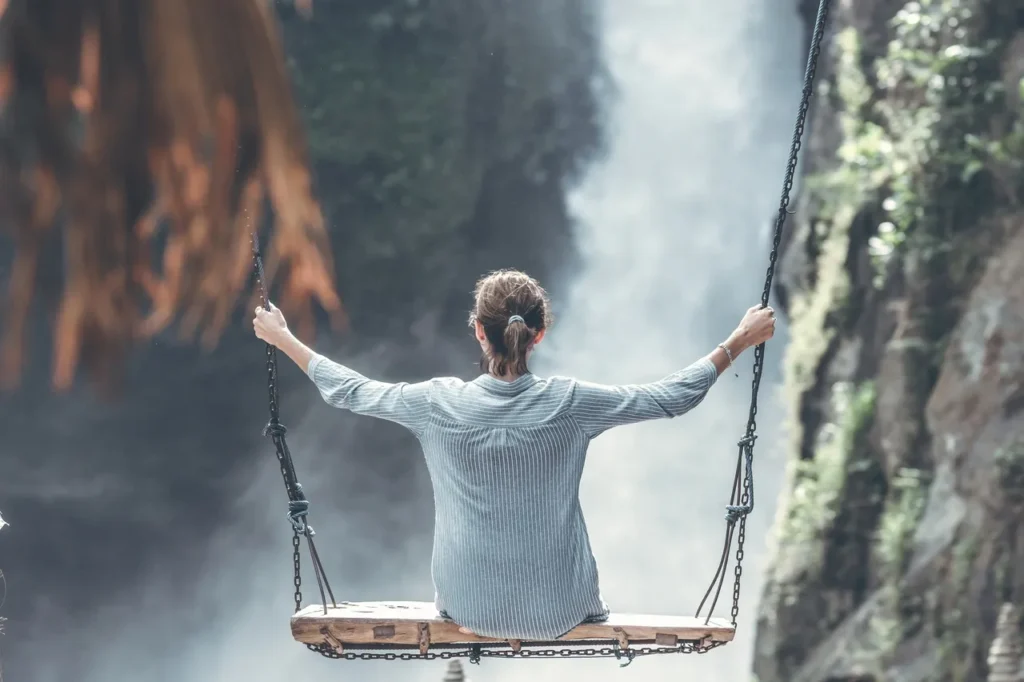 Woman sitting on a wooden swing with arms wide open, facing a waterfall in a natural canyon.