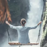 Woman sitting on a wooden swing with arms wide open, facing a waterfall in a natural canyon.