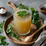 Glass of bone broth garnished with parsley, served on a wooden tray with herbs, salt, and pepper on the side.