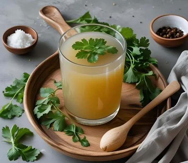 Glass of bone broth garnished with parsley, served on a wooden tray with herbs, salt, and pepper on the side.
