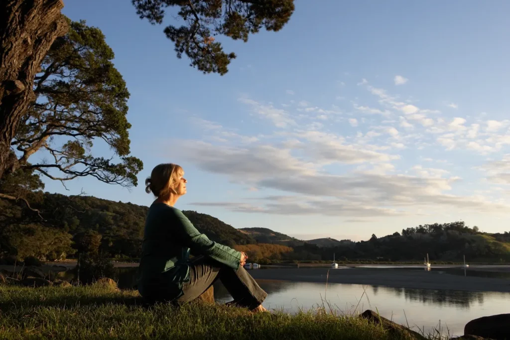 Woman sitting cross-legged on grass near a lake at sunset, surrounded by trees and calm water.